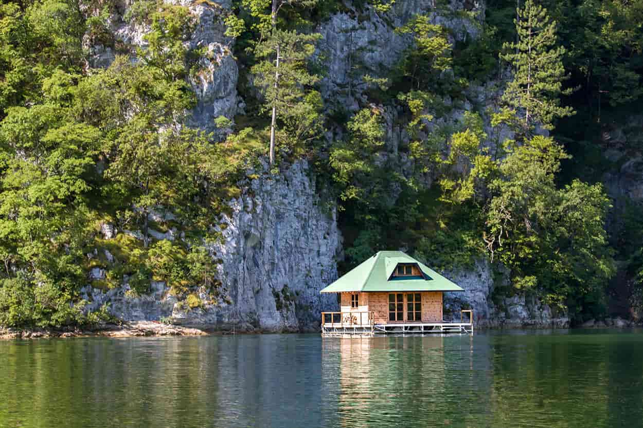 Drvena kućica na vodi na jezeru Perućac, okružena zelenim brdima.